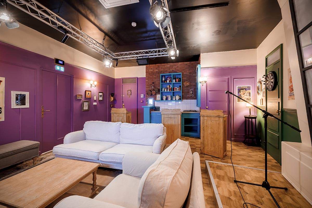 Photo 2 de l'intérieur du salon Sitcom12 (pour 12 personnes), avec une scène de karaoké, un plateau de quizz, des fauteuils et canapés, des fléchettes. Décors d'un plateau de tournage du salon d'une série TV