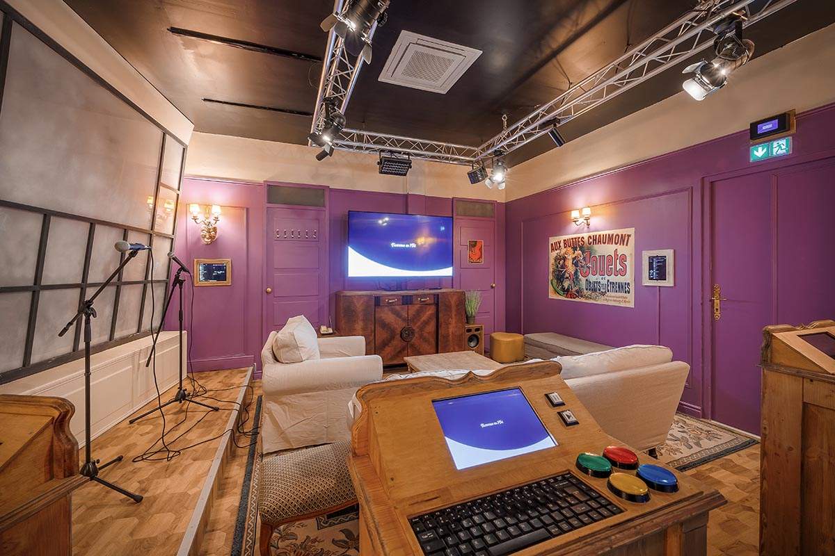 Photo 3 de l'intérieur du salon privatif Sitcom12 (pour 12 personnes), avec une scène de karaoké, un plateau de quizz, une TV, des fléchettes, des fauteuils et canapés. Décor d'un plateau de tournage d'une série TV