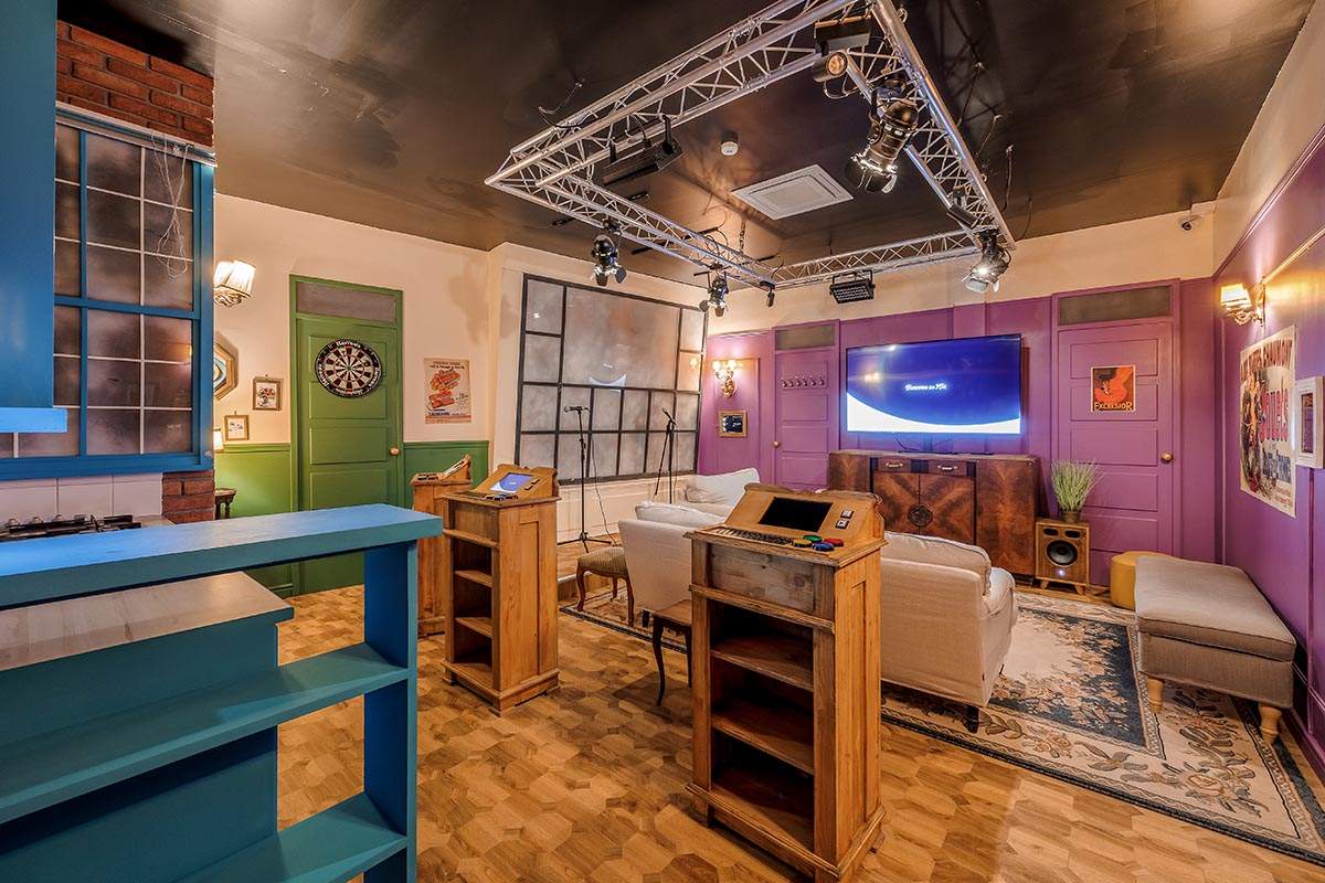 Photo 4 de l'intérieur du salon privatif Sitcom12 (pour 12 personnes), avec une scène de karaoké, un plateau de quizz, une TV, des fléchettes, des fauteuils et canapés. Décor d'un plateau de tournage d'une série TV