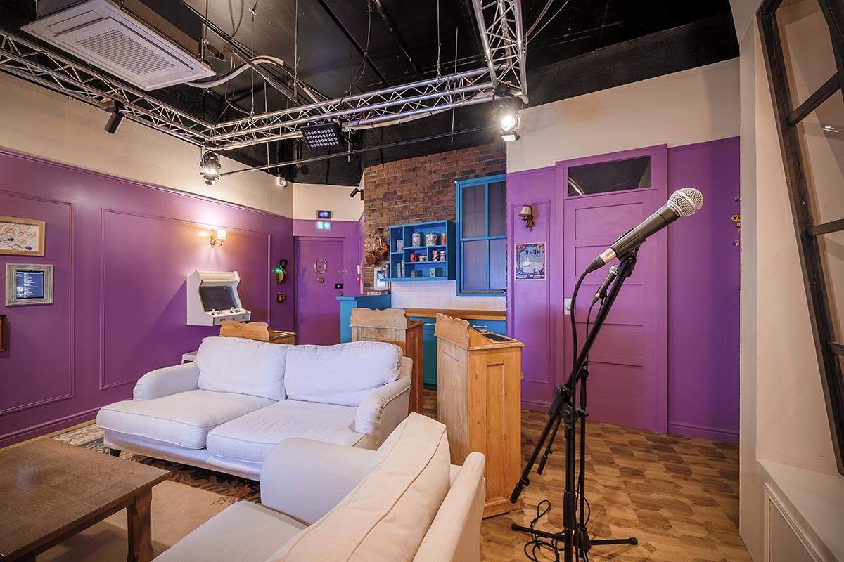 Photo 2 de l'intérieur du salon privatif Sitcom6 (pour 6 personnes), avec une scène de karaoké, un plateau de quizz, une TV, des fléchettes, des fauteuils et canapés. Décor d'un plateau de tournage d'une série TV