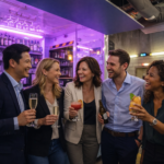 Photo d’un team-building au LE 3BIS : cinq adultes souriants, habillés en tenue chic décontractée, se tiennent debout autour du bar dans une ambiance chaleureuse aux lumières violettes. Ils lèvent leurs verres – cocktails colorés, flûtes de champagne et bière – en riant, dans un décor industriel moderne avec briques apparentes, étagères de bouteilles et éclairage néon, illustrant un moment convivial et festif entre collègues.