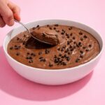 Bol de mousse au chocolat onctueuse parsemée de pépites de chocolat, dans lequel une cuillère vient prélever une portion, sur fond rose. 🍫🥄