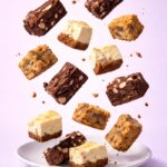 Assortiment de desserts de brunch composé de brownies aux noix, blondies et carrés cheesecake, disposés en suspension au-dessus d’une assiette sur fond pastel. 🍰🍫